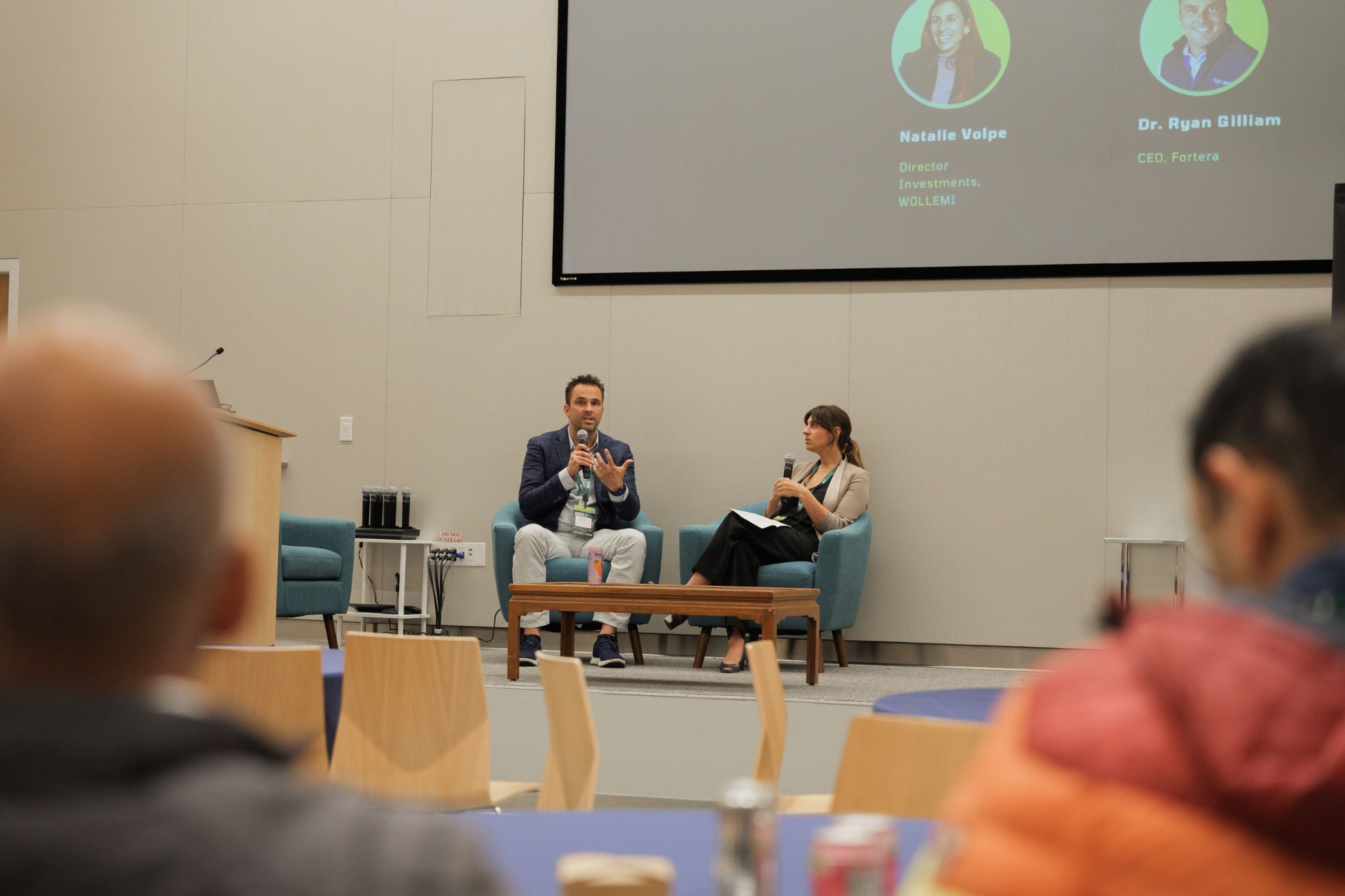 Capital Day fireside chat 'Crossing the Deployment Valley — Founder Perspective' viewed from the audience — Dr. Ryan Gilliam, CEO of Fortera, speaking with mic beside moderator Natalie Volpe of WOLLEMI on the UC Law SF stage