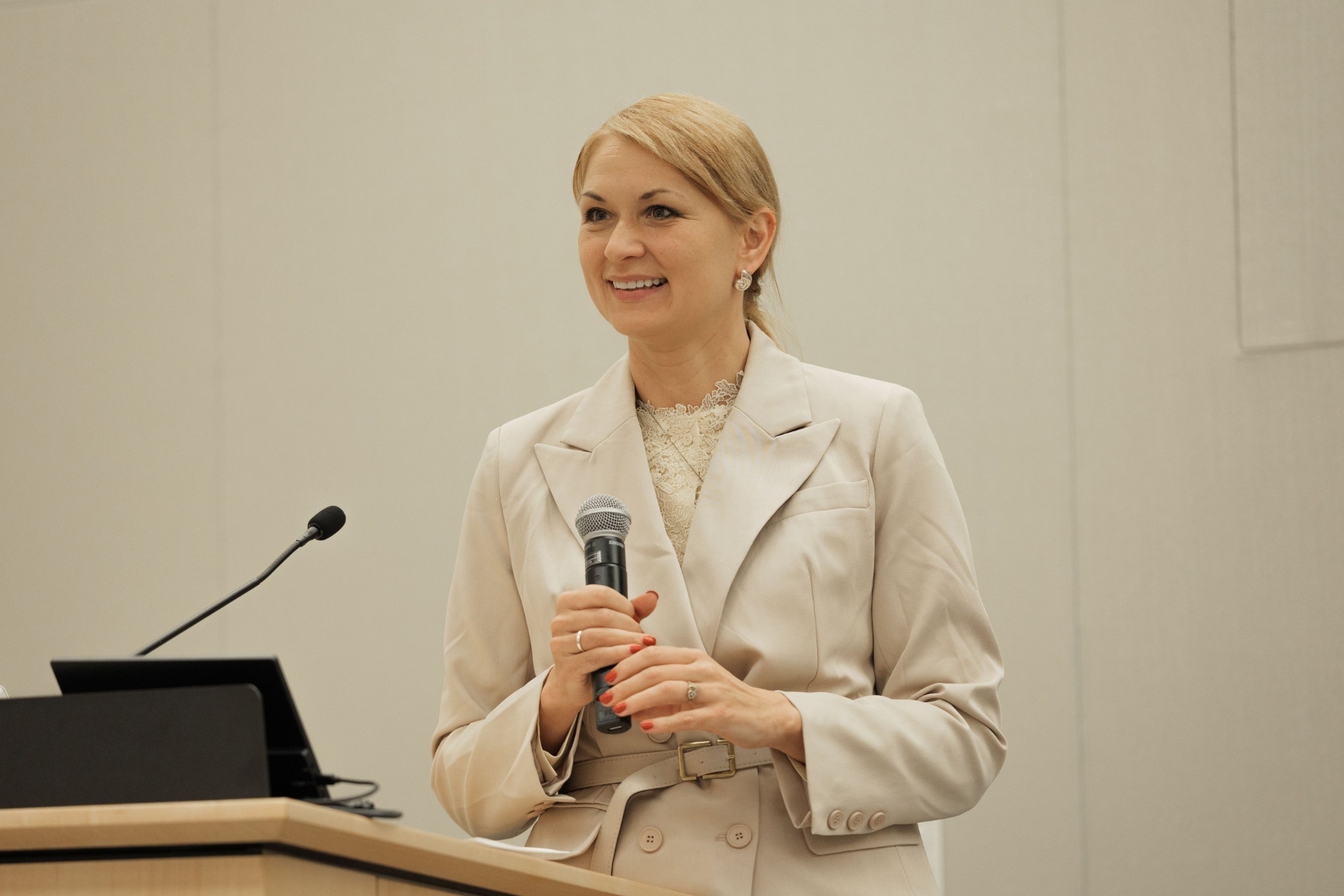 Portrait of a blonde speaker in a beige blazer smiling while holding a microphone at the UC Law SF podium during Capital Day