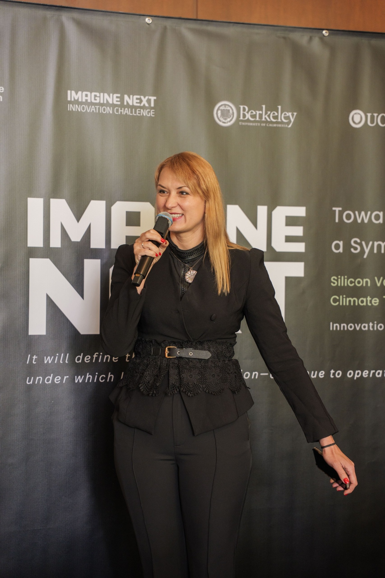 Host smiling at the podium beside the Imagine Next backdrop